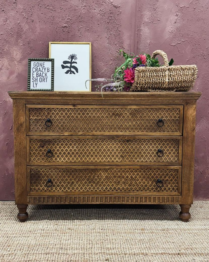A handcrafted carved mango wood chest of drawers with intricate diamond designs, polished finish, and three drawers visible. The chest is placed against a purple wall with a framed picture and a basket of flowers on top for decorative purposes.